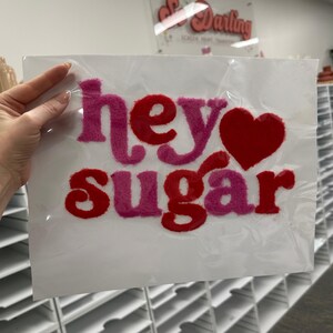 May include: A white canvas with the words "hey sugar" in pink and red, with a red heart replacing the "o". The letters have a fuzzy texture. The canvas is in a clear plastic sleeve.