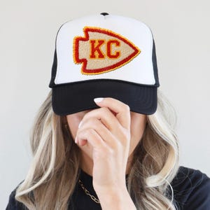 May include: A black and white trucker hat with a white front panel featuring a red and yellow arrowhead design. The arrowhead has the letters "KC" in the center. The hat has a black brim and mesh back.