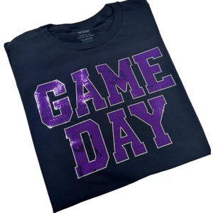 May include: A black short-sleeved t-shirt featuring the text GAME DAY in large, block-style letters. The letters are filled with a dense pattern of small, reflective purple rhinestones, creating a textured, shimmering appearance against the fabric.