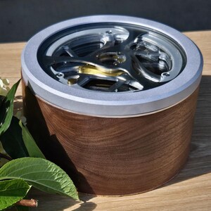 May include: A round wooden box with a silver rim and a clear top revealing intricate metal gears. The box has a dark brown wood grain finish. The gears are silver and gold. The box is sitting on a wooden surface.