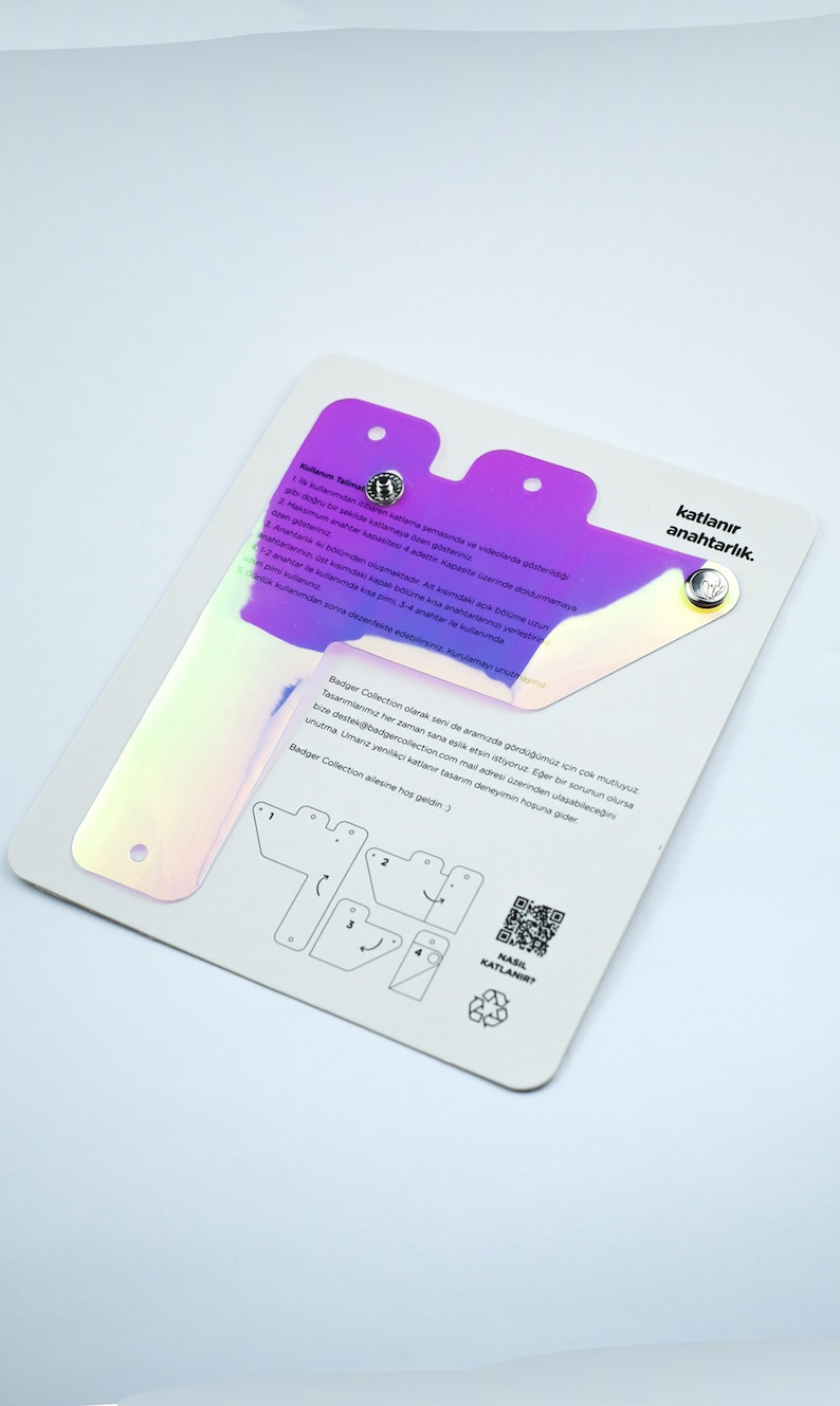 Puede incluir: Una tarjeta blanca con un dise&ntilde;o hologr&aacute;fico morado y rosa. La tarjeta tiene instrucciones sobre c&oacute;mo doblar un llavero, con un diagrama que muestra los pasos. La tarjeta tambi&eacute;n tiene un c&oacute;digo QR y el texto "katlanır anahtarlık." que se traduce como "llavero plegable."