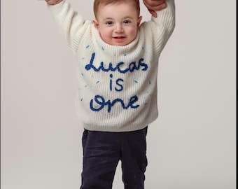 May include: A white knitted sweater with blue embroidered text that reads "Lucas is one". The sweater is being worn by a toddler.
