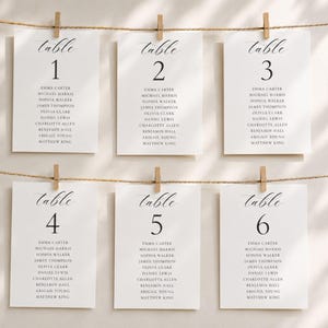 Könnte beinhalten: Sechs weiße, rechteckige Tischkarten für Hochzeiten, die an zwei horizontalen Schnüren mit Holzklammern hängen. Jede Karte zeigt das Wort table, gefolgt von einer Zahl von eins bis sechs und einer Liste von Namen in schwarzer Serifenschrift.