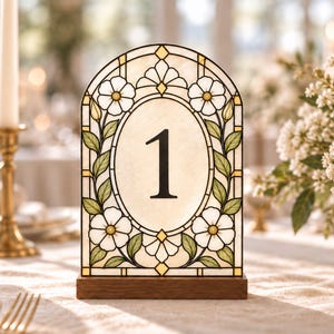 May include: An arched table number sign featuring the number 1 in a black serif font. The sign has a stained glass style design with white flowers, green leaves, and gold accents, set within a rectangular dark brown wooden base stand.