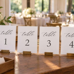 以下が含まれることがあります： Table 1、Table 2、Table 3、Table 4と黒の筆記体およびセリフ体で記された4枚の白い長方形のテーブルナンバーカード。各カードは、木製の表面に置かれた小さな長方形のゴールドトーンの金属製スタンドによって直立に保持されています。