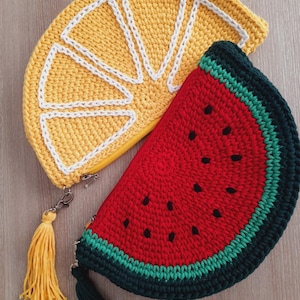 May include: Two crocheted purses shaped like fruit slices. One is a yellow lemon slice with white stitching and a yellow tassel. The other is a red watermelon slice with black seeds and a green tassel.