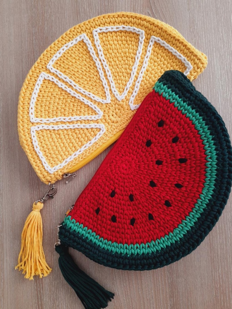 May include: Two crocheted purses shaped like fruit slices. One is a yellow lemon slice with white stitching and a yellow tassel. The other is a red watermelon slice with black seeds and a green tassel.