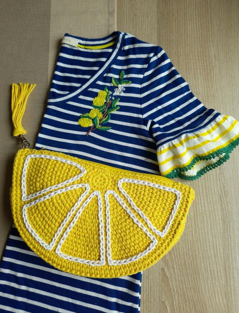 May include: A yellow crocheted lemon-shaped purse with a white tassel. The purse is sitting on a blue and white striped shirt with a lemon embroidery.