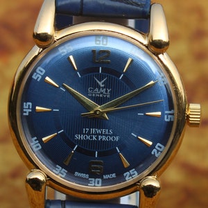 Może przedstawiać: Złoty zegarek z granatową tarczą i pasującym skórzanym paskiem. Tarcza zegarka ma złote wskazówki i znaczniki z napisem "CAMY GENEVE 17 JEWELS SHOCK PROOF". Pasek ma szerokość około 2 cm.