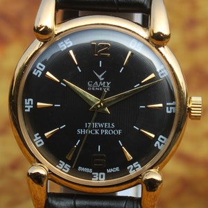 Peut inclure: Une montre vintage dorée avec un cadran noir et un bracelet en cuir noir. Le cadran de la montre comporte des aiguilles et des marqueurs dorés, avec le texte "CAMY GENEVE", "17 JEWELS" et "SHOCK PROOF". La montre a un cadran rond et un bracelet en cuir noir.
