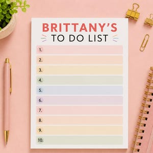 Peut inclure: Un bloc-notes rectangulaire avec le texte BRITTANY'S TO DO LIST en haut, présentant dix lignes numérotées dans des couleurs pastel alternées, notamment rose, orange, jaune, vert, bleu et violet, sur un fond rose clair uni.