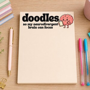May include: A rectangular notepad with a cream-coloured paper surface featuring the black text doodles so my neurodivergent brain can focus and a cartoon illustration of a pink brain character holding a clipboard and a pencil.