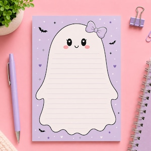 May include: A rectangular notepad with a light purple background featuring a central white ghost character with a lavender bow, pink cheeks, and heart-shaped eyes. The ghost contains horizontal lines for writing, surrounded by small bats, stars, and hearts.