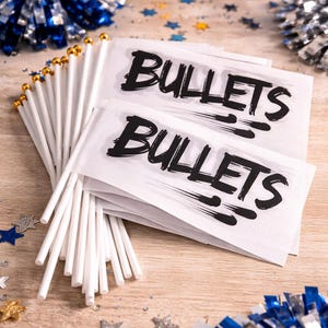Könnte beinhalten: Weiße Flaggen mit dem Wort "BULLETS" in schwarzer Schrift, mit goldfarbenen Stäben. Die Flaggen liegen auf einer Holzoberfläche, umgeben von blauen und goldenen Pompons und sternförmigem Konfetti. Im Hintergrund befindet sich ein blau-weißer Papierkegel.