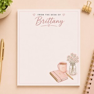 Peut inclure: Un bloc-notes rectangulaire avec une bordure rose pâle et le texte "FROM THE DESK OF Brittany" en haut. Le coin inférieur droit présente une illustration imprimée d'une tasse rose, d'un carnet à spirales rose, d'un stylo et d'un bocal en verre avec des fleurs.