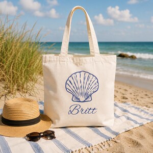 Peut inclure: Un sac fourre-tout en toile couleur crème orné d'un motif bleu représentant un coquillage et le prénom Britt écrit en lettres cursives bleues. Le sac possède deux anses et est posé près d'un chapeau de paille et de lunettes de soleil.