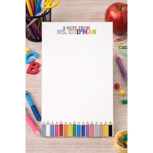 Puede incluir: Un bloc de notas blanco con el texto "A NOTE FROM MS. CHIPMAN" en letras coloridas. La parte inferior del bloc de notas presenta una fila de lápices de colores. Una manzana, lápices y otros útiles escolares rodean el bloc de notas.