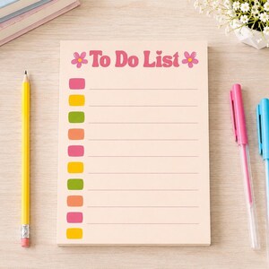 Peut inclure: Un bloc-notes rectangulaire avec le texte To Do List en rose, encadré par deux icônes de fleurs roses. La page comporte dix lignes horizontales, chacune précédée d'un petit rectangle coloré alternant entre le rose, le jaune, le vert et l'orange.