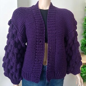 Op de afbeelding: Een paarse gebreide cardigan met een losse pasvorm en gepofte mouwen. De cardigan heeft een V-hals en een eenvoudig ontwerp.