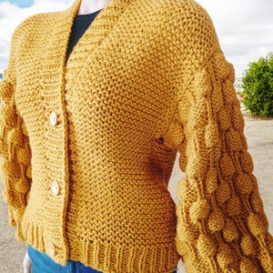 Op de afbeelding: Een mosterdgele cardigan met knopen en textuur in de mouwen. De cardigan is gebreid en heeft een ontspannen pasvorm.