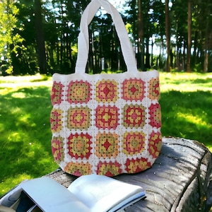 May include: A white crocheted tote bag with a colorful geometric pattern. The bag has a square shape and is made of a soft, textured yarn. The bag is sitting on a wooden surface.