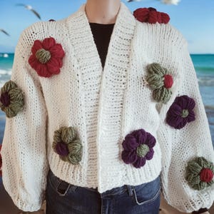 May include: White knitted cardigan with a V-neck and large, colorful flower embellishments. The cardigan has long sleeves and a relaxed fit.