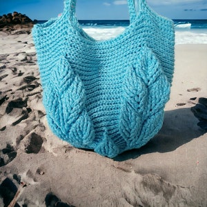 May include: A light blue crocheted tote bag with a leaf pattern. The bag is sitting on a sandy beach.