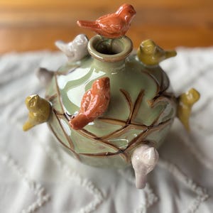May include: A ceramic decorative jar with a pale green glaze and brown detailing. The jar is decorated with several small, colourful bird figurines in shades of orange, yellow, and white. The jar sits on a white, textured surface.