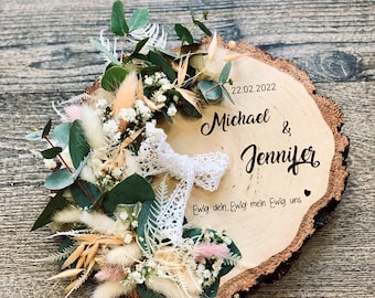 May include: A wooden slice with a white lace ribbon and dried flowers. The wood is engraved with the date "22.02.2022", the names "Michael & Jennifer", and the text "Ewig dein Ewig mein Ewig uns".
