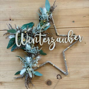 Könnte beinhalten: Ein rustikaler sternförmiger Kranz aus Schnur mit einem Holzschild, auf dem "Winterzauber" steht und der mit getrockneten Blumen und Grünpflanzen verziert ist.