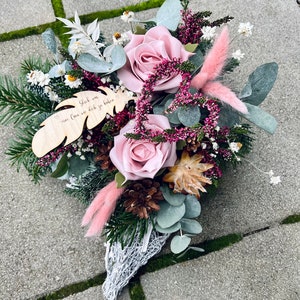 May include: A pink and white floral arrangement in a white wire basket. The arrangement includes pink roses, white flowers, and green foliage. A wooden sign with the text "Glück war, dass wir dich ja hatten" is included in the arrangement.