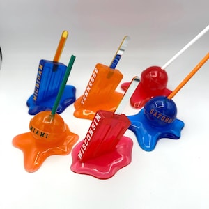 May include: Five colorful melted popsicle holders with different college logos. The logos are "Michigan", "Tennessee", "Wisconsin", "Miami", and "Gators".