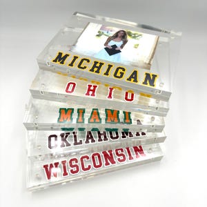 May include: Clear acrylic photo frames stacked, each with a different university name in bold lettering: Michigan, Ohio, Miami, Oklahoma, and Wisconsin. A photo of a person in a graduation gown is visible in the top frame.