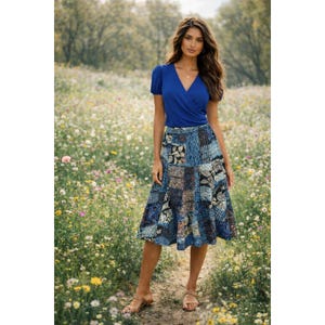 May include: A woman wearing a blue wrap top and a patchwork skirt. The skirt features various blue patterns and designs. She is wearing sandals and standing in a field of wildflowers.