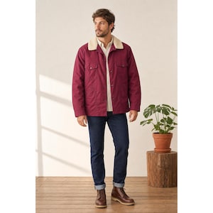 May include: A burgundy jacket with a cream-colored shearling collar, worn over a light-colored button-down shirt. The jacket has two chest pockets and is paired with dark blue jeans and brown leather boots. A potted plant sits on a wooden stand.
