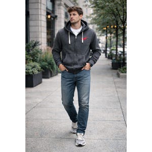 May include: A man wearing a dark gray zip-up hoodie with a red logo on the chest, a white t-shirt, and blue jeans. He is walking on a sidewalk, wearing gray and white sneakers. The hoodie has a hood with white drawstrings.
