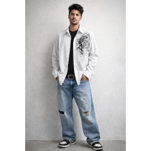 May include: A white button-down shirt with a black floral design on the front, paired with a black t-shirt, light blue ripped jeans, and black and white sneakers. The outfit includes a black belt with a silver chain.