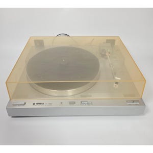 Może przedstawiać: Srebrny gramofon Yamaha P-700 z przezroczystą, bursztynową osłoną przeciwpyłową. Gramofon posiada srebrny talerz, ramię i przyciski sterowania. Logo i numer modelu Yamaha są widoczne.