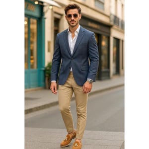 May include: A man wearing a navy blue blazer over a white dress shirt, paired with khaki pants and brown loafers. The outfit is completed with sunglasses and a silver watch. The setting is an outdoor urban environment.