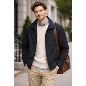 May include: A person wearing a navy blue jacket over a cream-colored cable-knit sweater and a blue and white checkered shirt. The person is also wearing khaki pants and carrying a brown leather shoulder bag.