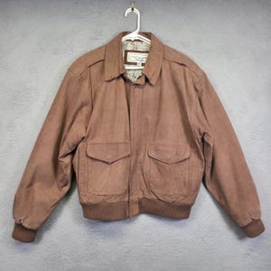 May include: A brown leather bomber jacket with two front pockets and a ribbed hem and cuffs. The jacket is hanging on a white hanger against a gray background. The jacket has a collared neckline and a label that reads "Luckman".