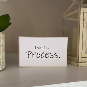 May include: White wooden sign with black text that reads "Trust The Process."