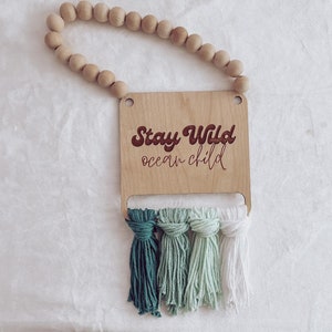 May include: Wooden sign with the text "Stay Wild ocean child" in a retro font. The sign has three green and white tassels hanging from it. The sign is hanging from a wooden bead garland.