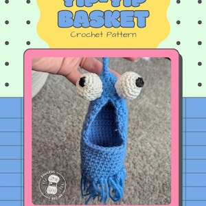 Può includere: Un cestino blu all'uncinetto con grandi occhi bianchi e neri, simile a un personaggio dei cartoni animati. Il testo "YIP-YIP BASKET Crochet Pattern" è visualizzato sopra il cestino. L'immagine è incorniciata in rosa e su uno sfondo blu e verde.