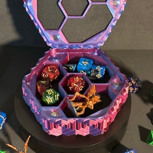 May include: A hexagonal dice box with a purple and pink iridescent finish. The box has a removable lid and six compartments for dice. The box is filled with dice and a miniature figurine.