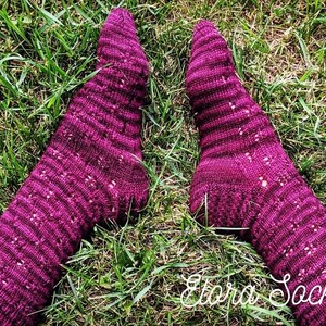 May include: A pair of purple knitted socks with a lacy pattern. The socks are on a bed of green grass.