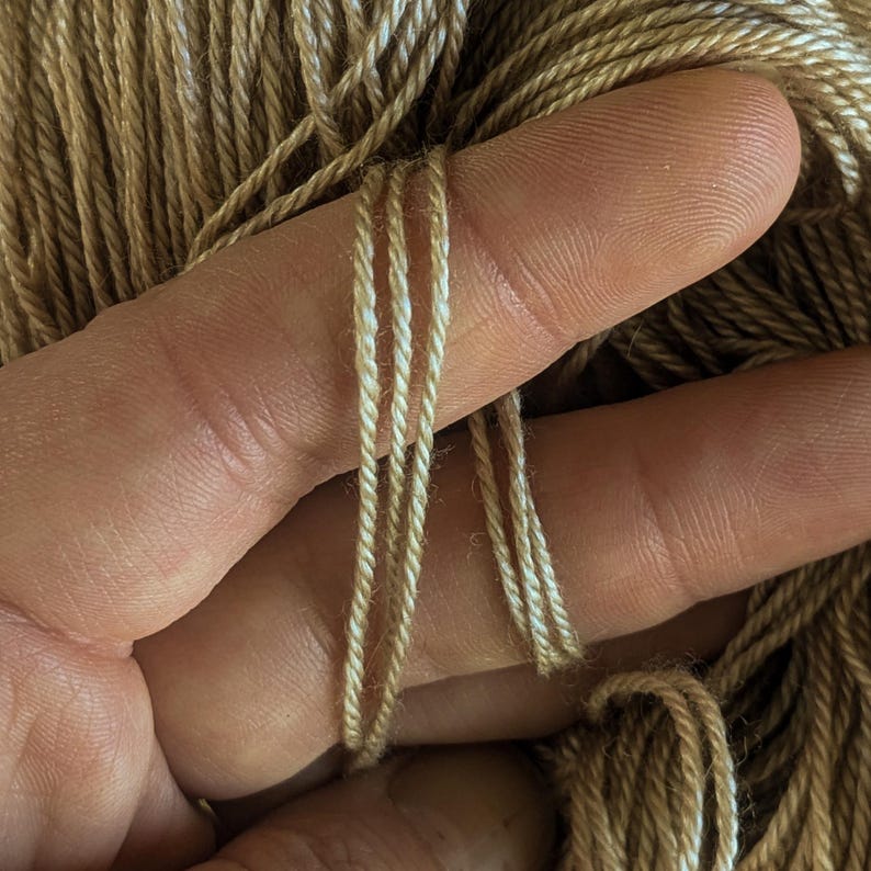 May include: Close-up of a hand holding a bundle of brown and white yarn. The yarn is soft and fluffy, and the colours are subtle and natural.