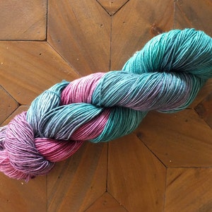 May include: A skein of yarn in shades of teal, purple, and pink. The yarn is twisted and has a gradient color effect.