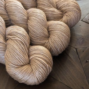 May include: Three skeins of beige yarn, wound into balls, on a wooden surface.