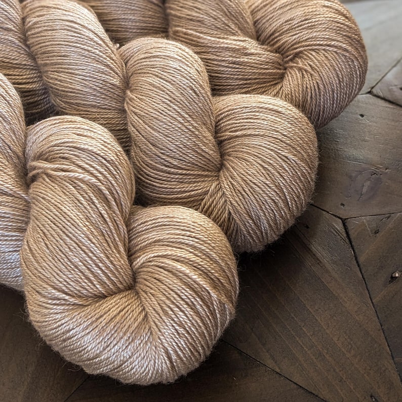 May include: Three skeins of beige yarn, wound into balls, on a wooden surface.